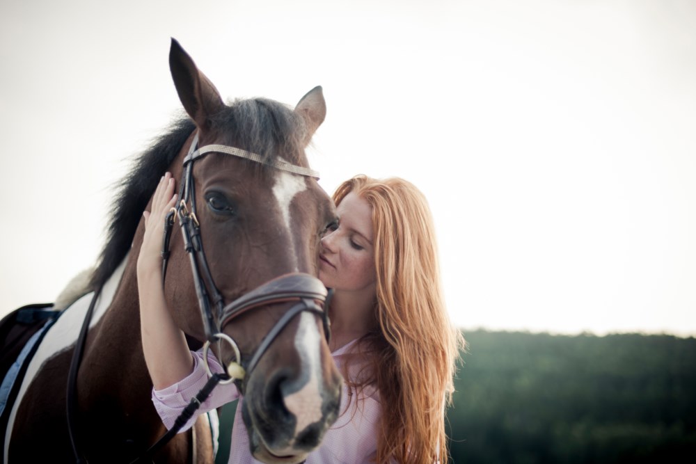 En kvinne med rødt hår lukker øynene og hviler sitt kinn mot en hest sitt hode. Hesten, med en hvit blis og utstyrt med et svart hodelag, ser mot kameraet. Bildet er tatt utendørs med en lys, uskarp bakgrunn som gir en rolig og fredelig atmosfære.